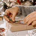 Person verpackt Weihnachtsgeschenk mit braunem Papier und Kordel, umgeben von Deko, Lichtern und Kaffeetasse.