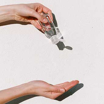 Eine Hand mit kleiner Flasche Desinfektionsmittel tropft Inhalt auf eine darunterliegende Hand vor grauen Hintergrund.