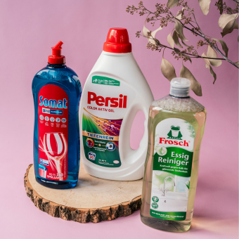 Somat Spülmittel, Persil Waschmittel und Frosch Essig Reiniger auf Holzscheibe vor rosa Hintergrund präsentiert.