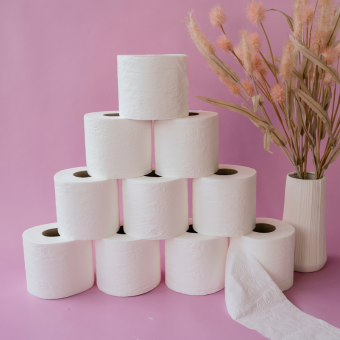 Neun weiße Toilettenpapierrollen als Pyramide auf rosa Hintergrund mit Vase und getrocknetem Pampasgras.