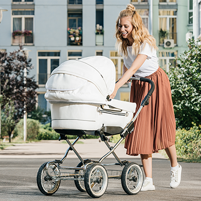 Frau schiebt weißen Kinderwagen auf Straße vor Wohnhaus.