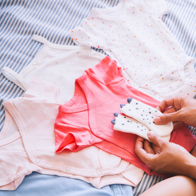 Person faltet Babykleidung auf gestreiftem Stoff – rosa Strampler, gemusterte Bodys, Babysocken, Erstausstattung.