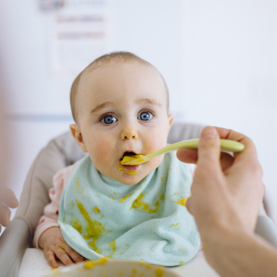Baby im Hochstuhl mit verschmiertem Lätzchen wird gefüttert – Fokus auf Beikost, Babyernährung und Familienalltag.