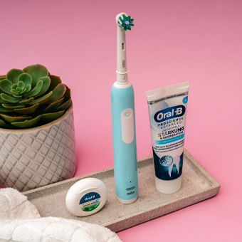 Oral-B Zahnbürste, Zahnseide und Zahnpasta auf Tablett – moderne Zahnpflege vor rosa Hintergrund.
