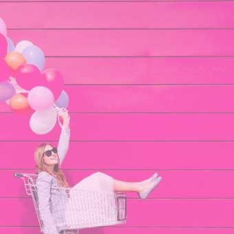 Frau mit bunten Luftballons sitzt im Einkaufswagen vor pinker Wand.