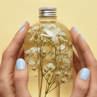 Hände mit hellblauem Nagellack halten Glasflasche mit weißen Blüten vor gelbem Hintergrund.
