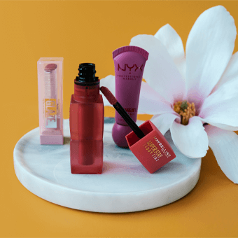 NYX Lipgloss, Maybelline Lippenstift & Lip Balm auf Marmor-Tablett mit Blume – Beauty-Produkte im Sommer-Look.