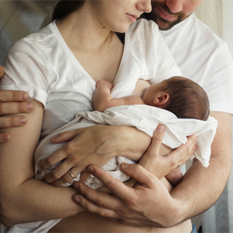 Mutter und Vater mit Baby im Arm. Mutter stillt das Baby und der Vater unterstützt sie dabei. 