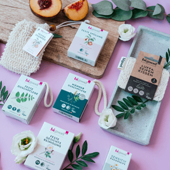 Naturkosmetik auf rosa Fläche mit Blüten, Blättern & Pfirsich – Seife, Duschgel, Gesichtsreiniger, vegane Pflege.