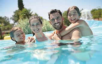 Familie entspannt im Pool an sonnigem Tag, umgeben von Natur und Wasserfall im Hintergrund.