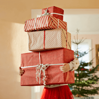 Person trägt gestapelte Weihnachtsgeschenke durch Flur, im Hintergrund festlich geschmückter Christbaum.