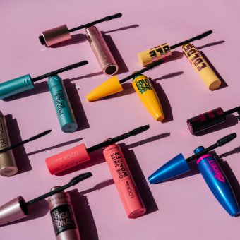 Mehrere bunte Mascara-Tuben mit Bürsten, flach auf rosa Hintergrund arrangiert – verschiedene Marken & Farben.