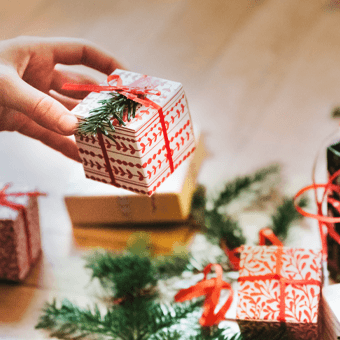 Hand hält kleines Weihnachtsgeschenk mit roter Schleife und Tannenzweig, weitere Päckchen auf Holztisch.