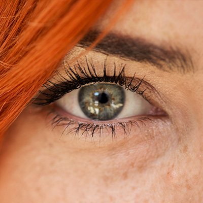 Nahaufnahme eines Auges mit grauer Iris, langen Wimpern, orangefarbenem Haar und Sommersprossen