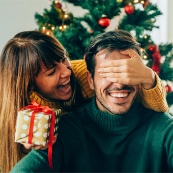 Zwei Personen in festlicher Umgebung, eine mit Rentiergeweih-Stirnband, Weihnachtsdeko und Geschenk mit roter Schleife.
