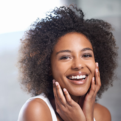 „Person mit lockigem Haar in weißem Oberteil, berührt mit beiden Händen das Gesicht