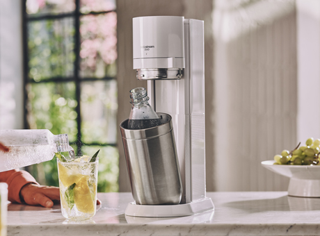 SodaStream Gerät auf Marmorplatte mit Flasche – frisches Getränk mit Eis, Limette und Minze, Trauben im Hintergrund