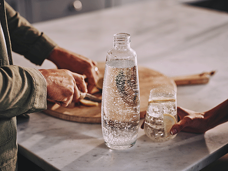 Zubereitung eines Erfrischungsgetränks mit Zitrone und Sprudelwasser in der Küche – Hände, Schneidebrett, Glas und Flasche