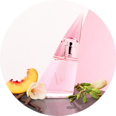 Parfümflasche mit rosa Flüssigkeit auf grauem Hintergrund, dekoriert mit Pfirsichscheibe, Blüten und grünen Blättern