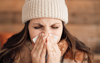 Person trägt warme Strickmütze und Schal, hält Hände vor Mund, Fokus auf Schutz vor Kälte und Erkältung im Winter.