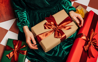 Person in grünem Samtkleid hält festlich verpacktes Geschenk mit roter Schleife, weitere Geschenke auf kariertem Boden.