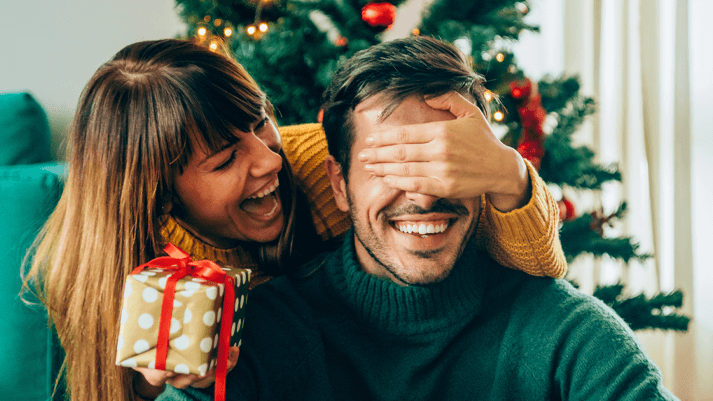 Paar sitzt vor geschmücktem Weihnachtsbaum und hält Geschenk mit roter Schleife – festliche Geschenkidee.