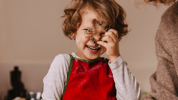 Kind mit Locken in roter Schürze neben Erwachsenem in braunem Pullover – gemeinsames Backen zur Weihnachtszeit.