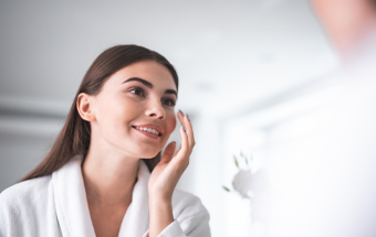 Person im weißen Bademantel betrachtet Haut im Spiegel, Fokus auf Gesichtspflege bei fettiger Haut.