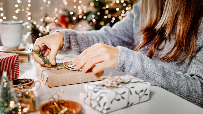 Person verpackt Geschenk mit braunem Papier und Zweig, im Hintergrund weitere Päckchen und Weihnachtsdeko.