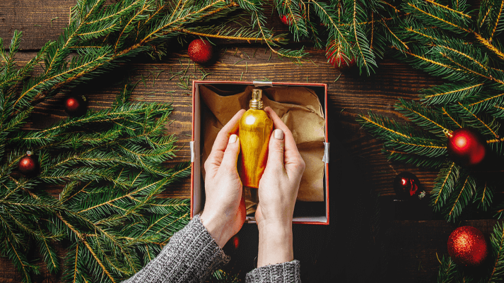 Hände halten goldfarbenes Parfumfläschchen in Geschenkbox, umgeben von Weihnachtsdeko und Tannenzweigen.