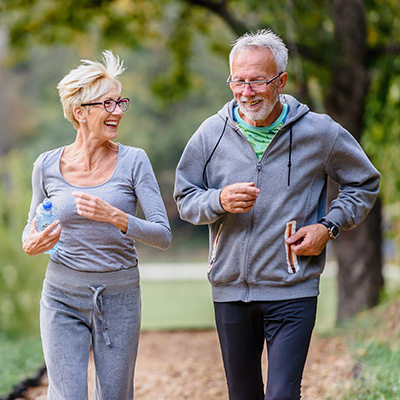 Zwei Personen joggen im Park in Sportkleidung mit Wasserflaschen, umgeben von Bäumen und einem Weg.