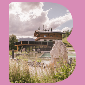 Alpines Hotel mit Naturteich und Bergkulisse, eingebettet in sommerliche Landschaft innerhalb eines pinken B‑Rahmens