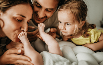 Familie mit Kleinkind in gelbem Shirt hält sanft die Füße eines Babys auf weißem Kissen – Geschwisterliebe, Familie, Neugeborenes. Copyright: Getty Images