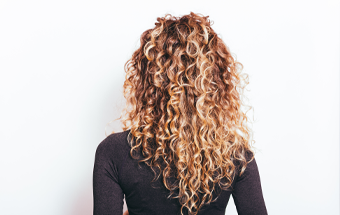 Rückansicht von Person mit langen, definierten Locken in Braun-Blond vor weißem Hintergrund, Fokus auf Lockenpflege.