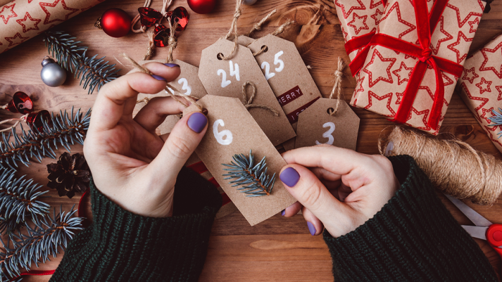 Hand hält Adventszahlen auf braunen Geschenkanhängern mit Geschenkbox, Tannenzweigen und Deko im Hintergrund.