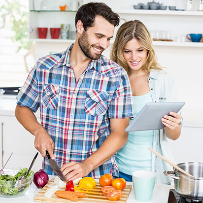 Mann schneidet Gemüse in moderner Küche, Frau daneben hält Tablet und schaut darauf