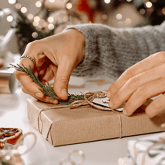 Person verpackt Weihnachtsgeschenk mit braunem Papier und Kordel, umgeben von Deko, Lichtern und Kaffeetasse.