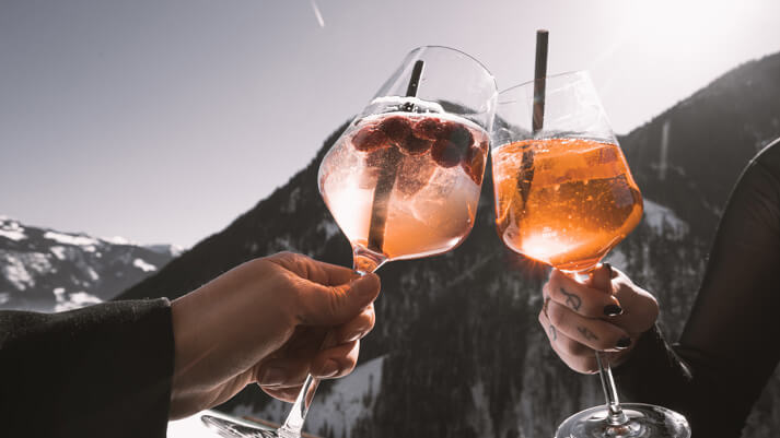 Zwei Personen stoßen mit Aperitif-Gläsern vor winterlicher Bergkulisse an; Genussmoment im alpinen Outdoor‑Ambiente.