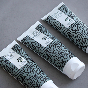 Drei Tuben Australian Bodycare Körperpflege mit Teebaumöl, blattähnlichem Muster auf grauem Hintergrund. - Link zu Markenseite/Körperpflege