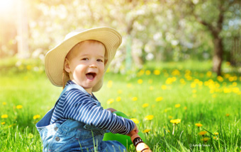 Kind mit Strohhut sitzt auf blühender Frühlingswiese mit gelben Blumen und Bäumen im Hintergrund. Copyright: Adobe Stock