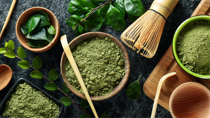 Matcha‑Pulver und Teezubehör mit frischen Teeblättern auf dunkler Oberfläche, perfekt für Superfood‑ und Tee‑Rezepte.