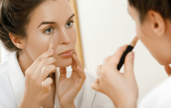 Person trägt Concealer mit Pinsel vor Spiegel auf - Link zum Blogartikel Concealer auftragen, Tipps für jeden Hautton