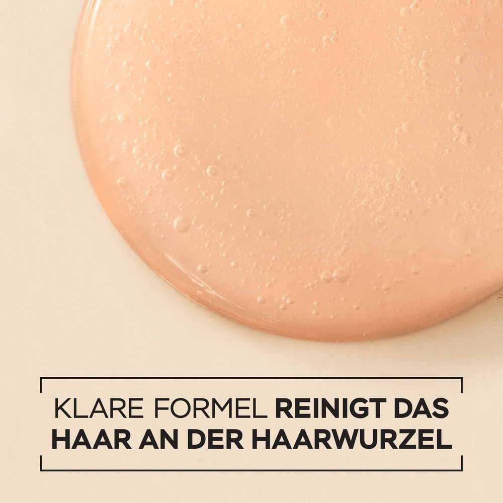 Nahaufnahme eines klaren, leicht bernsteinfarbenen Serums, das in einem glänzenden Tropfen auf einer beigefarbenen Oberfläche liegt und kleine Bläschen enthält. Darunter steht in schwarzer Schrift: KLARE FORMEL REINIGT DAS HAAR AN DER HAARWURZEL.