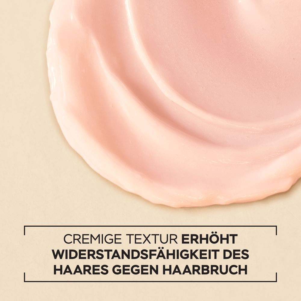 Nahaufnahme einer cremigen, rosafarbenen Textur, die wellenförmig auf einer beigefarbenen Oberfläche verteilt ist. Darunter steht in schwarzer Schrift: CREMIGE TEXTUR ERHÖHT WIDERSTANDSFÄHIGKEIT DES HAARES GEGEN HAARBRUCH.