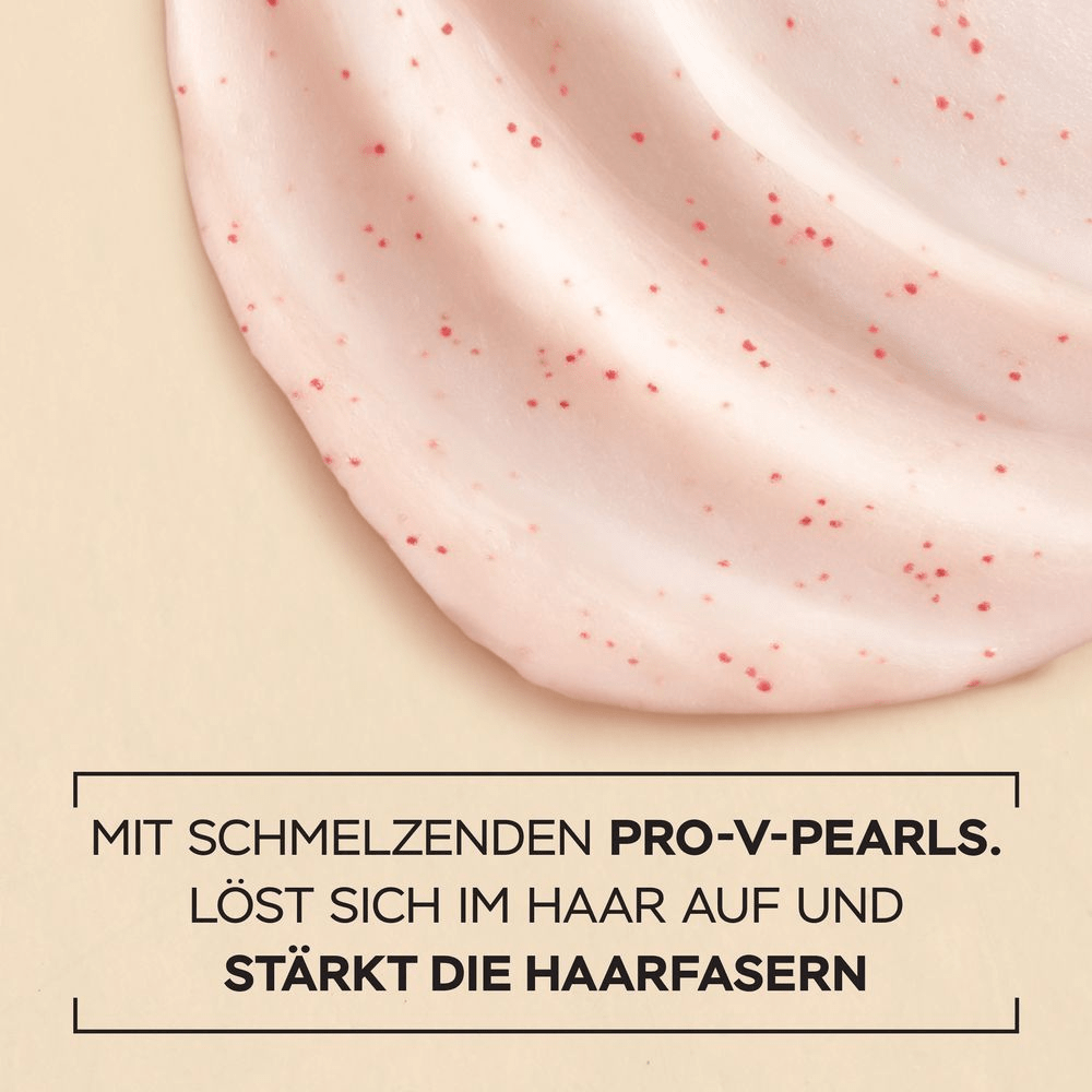 Nahaufnahme einer cremigen, rosafarbenen Textur mit kleinen roten Punkten (Perlen), die wellenförmig auf einer beigefarbenen Oberfläche verteilt ist. Darunter steht in schwarzer Schrift: MIT SCHMELZENDEN PRO-V-PEARLS. LÖST SICH IM HAAR AUF UND STÄRKT DIE HAARFASERN.