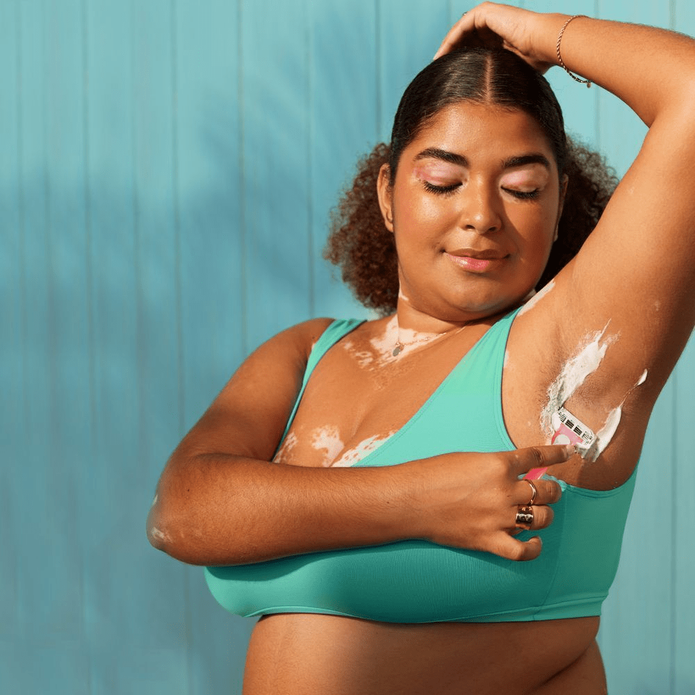 A woman with vitiligo smiles with her eyes closed while shaving her underarm with a pink razor, wearing a teal top against a light blue background.