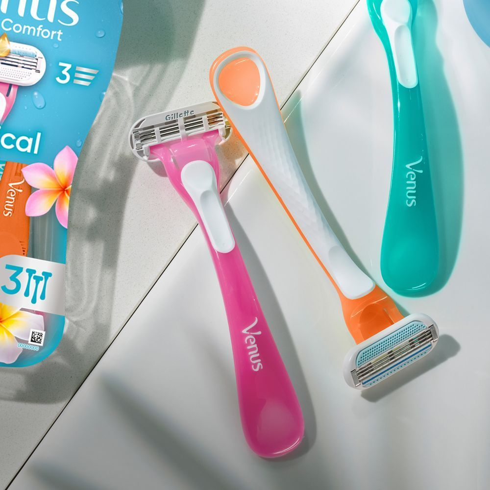 Three colorful Gillette Venus Comfortglide Tropical razors—pink, orange, and teal—are arranged on a white surface near the product packaging.