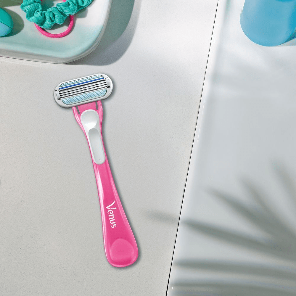 A pink Gillette Venus razor lies on a white bathroom counter next to a tray with colorful hair ties.