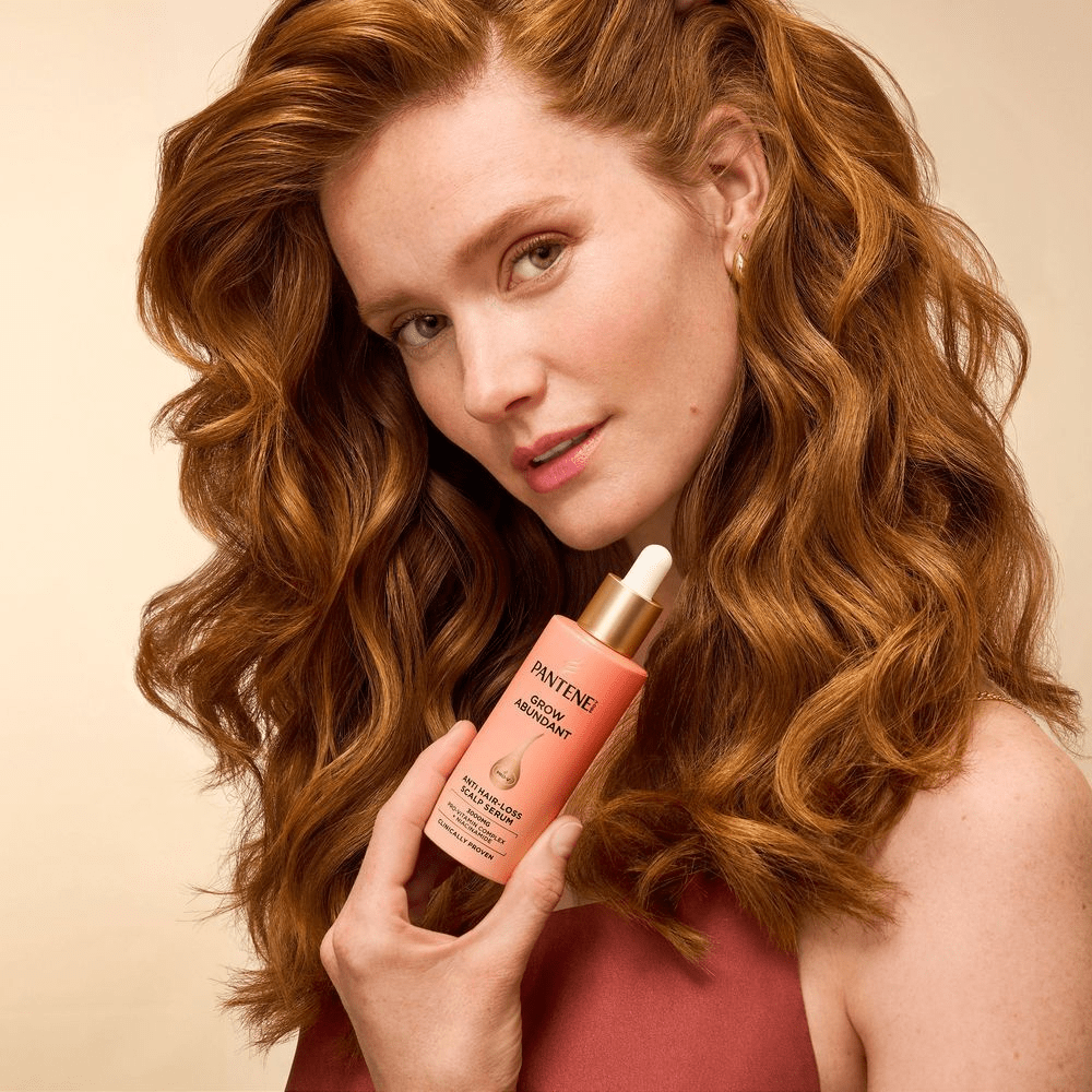 Eine Frau mit leuchtend roten, gewellten Haaren und hellem Teint hält lächelnd eine Pantene Pro-V Grow Abundant Anti-Haarverlust Kopfhautserumflasche in der Hand und blickt direkt in die Kamera. Ihr Haar fällt über ihre Schultern und ihr Oberteil ist bordeauxrot. Der Hintergrund ist beige.